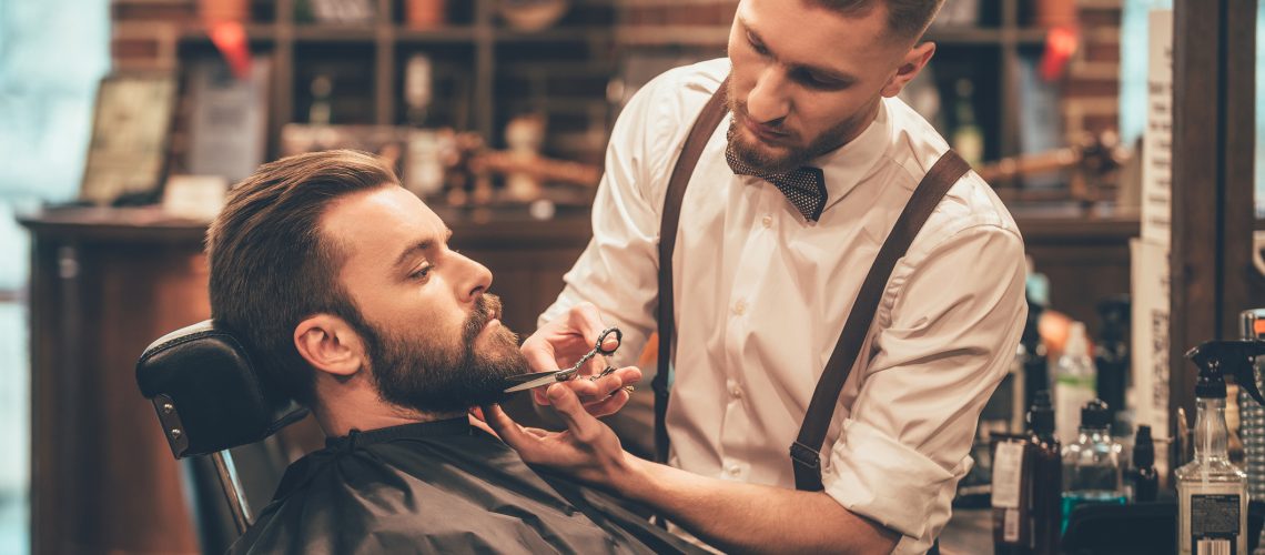 Side view of young bearded man getting beard haircut by hairdresser while sitting in chair at barbershop